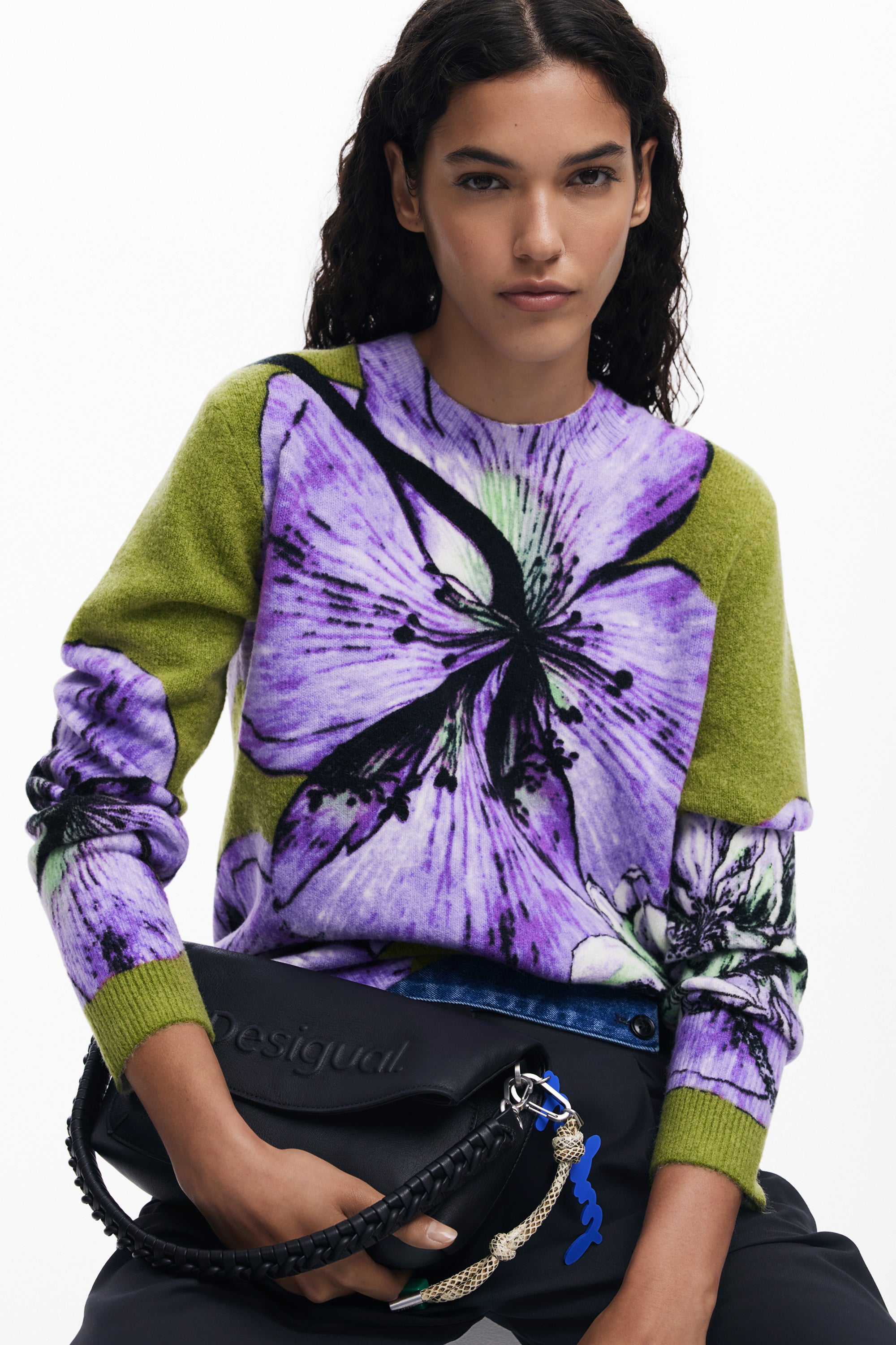 woman wearing a green and purple floral print jumper holding a black handbag on a white background