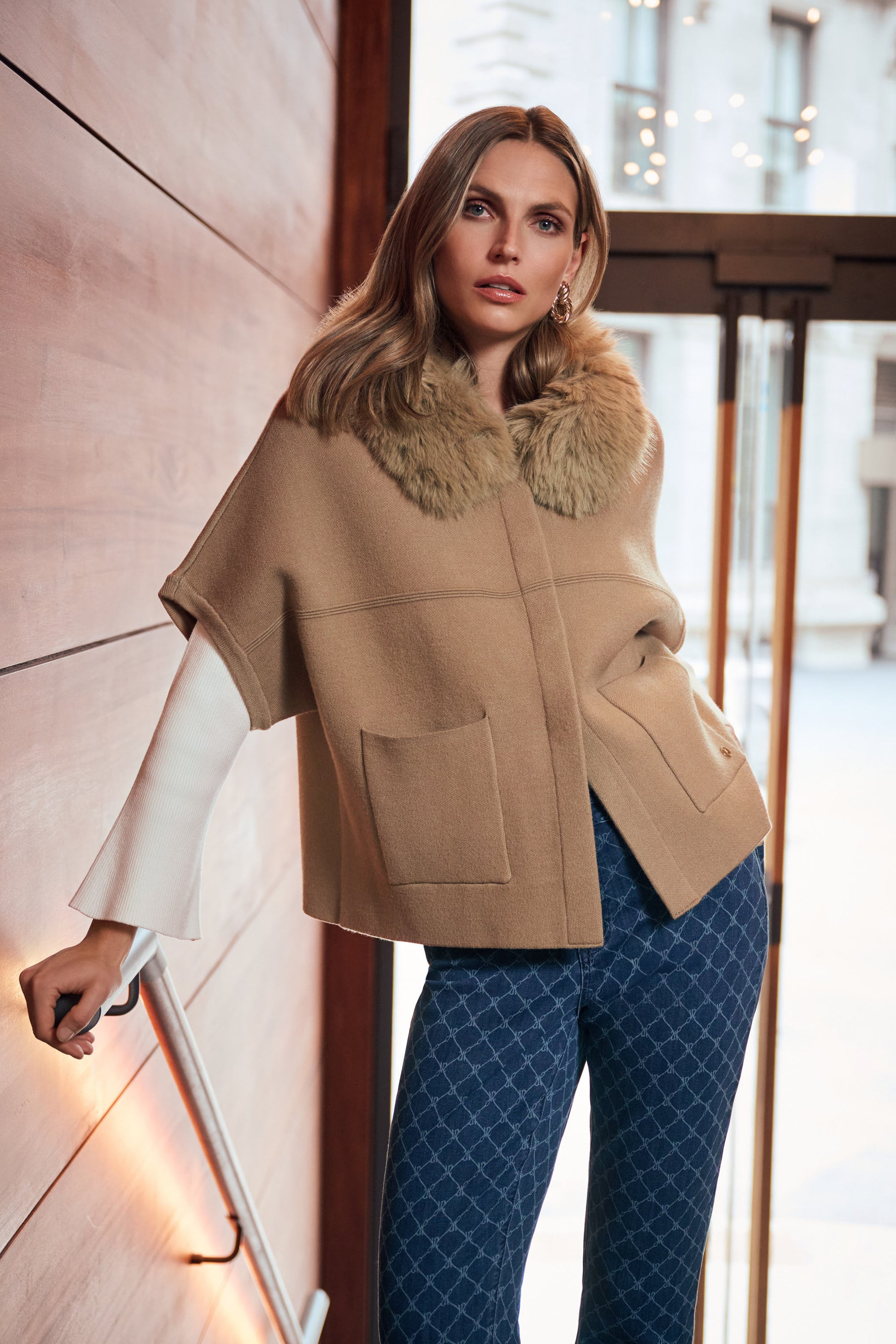 Model leaning against stone wall wearing beige cape jacket with fur collar Australia