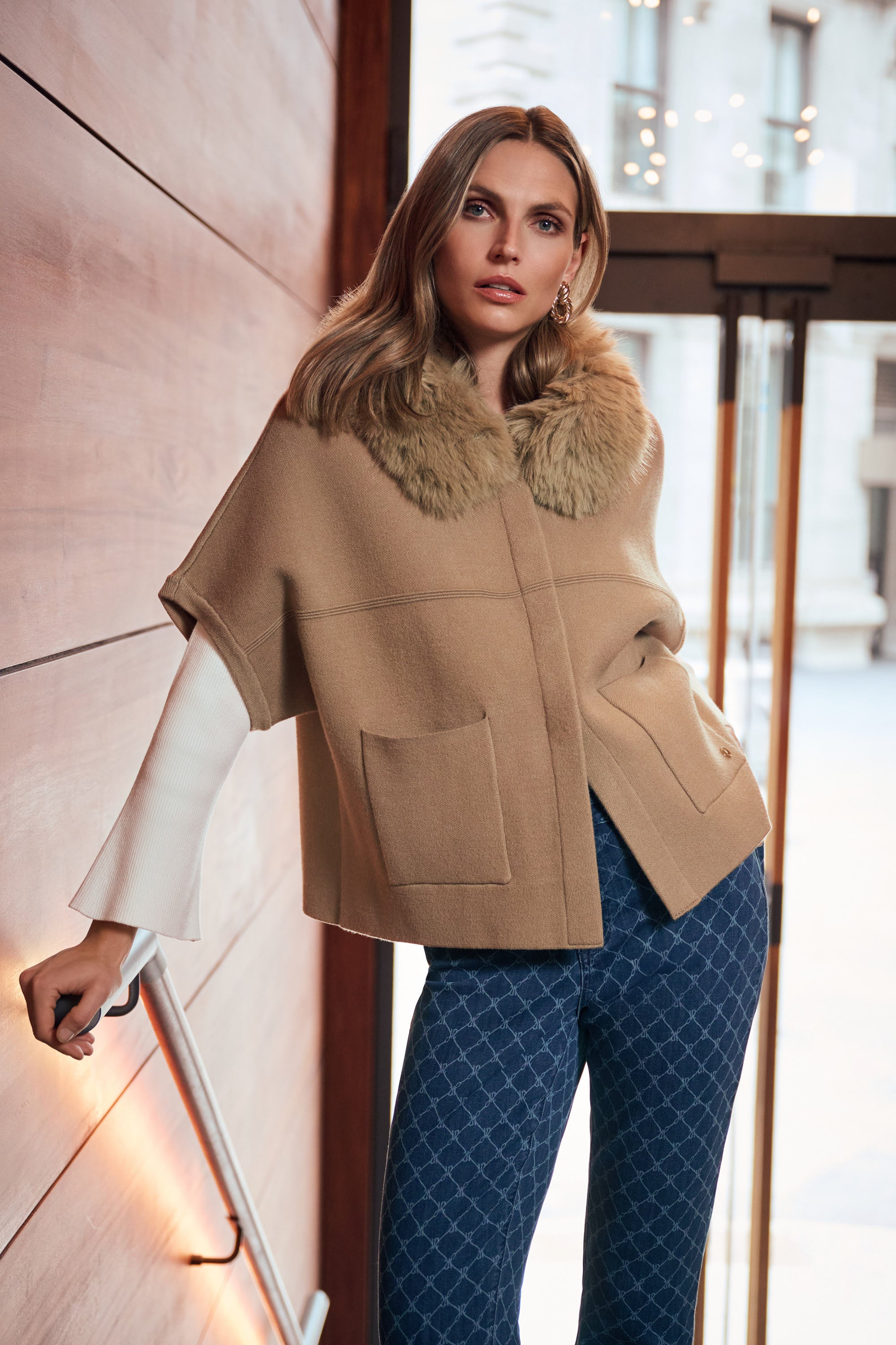 Model leaning against stone wall wearing beige cape jacket with fur collar Australia