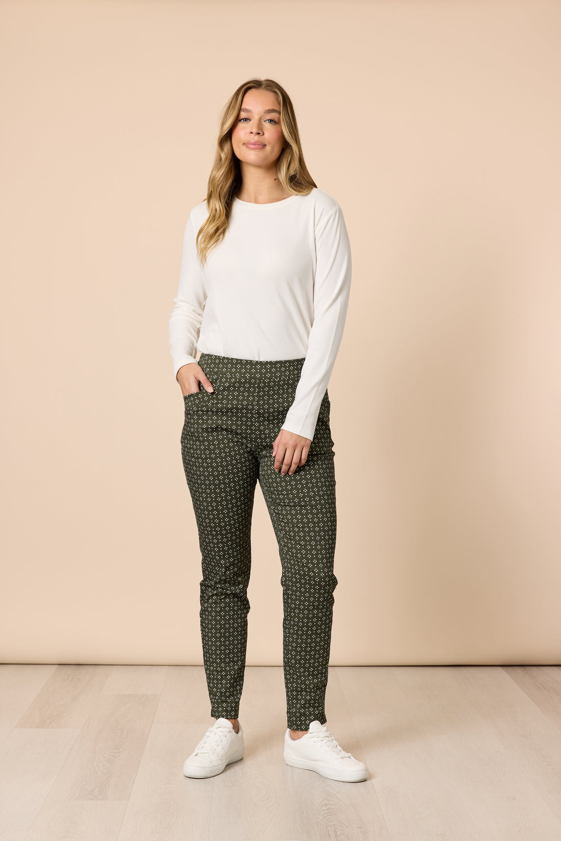 Woman wearing a classic ivory long sleeve basic top with olive printed pants and white sneakers, full body casual outfit on a beige background Showies Boutique