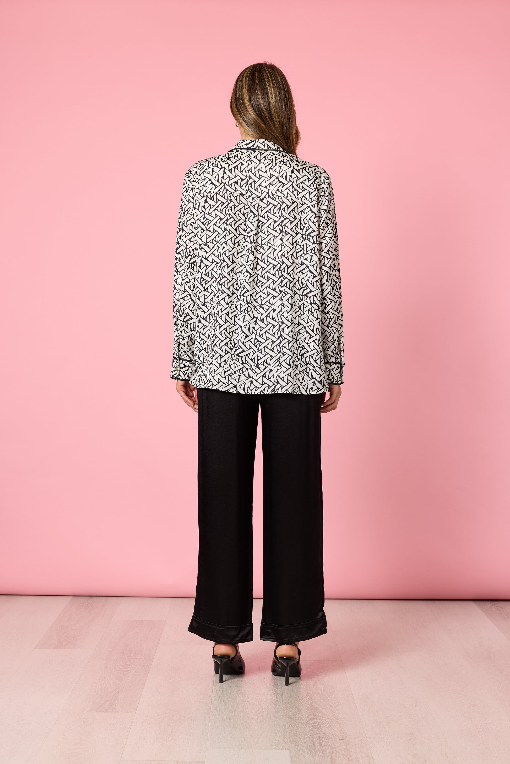 Back view of woman in black and ivory print blouse with loose black pants on pink background