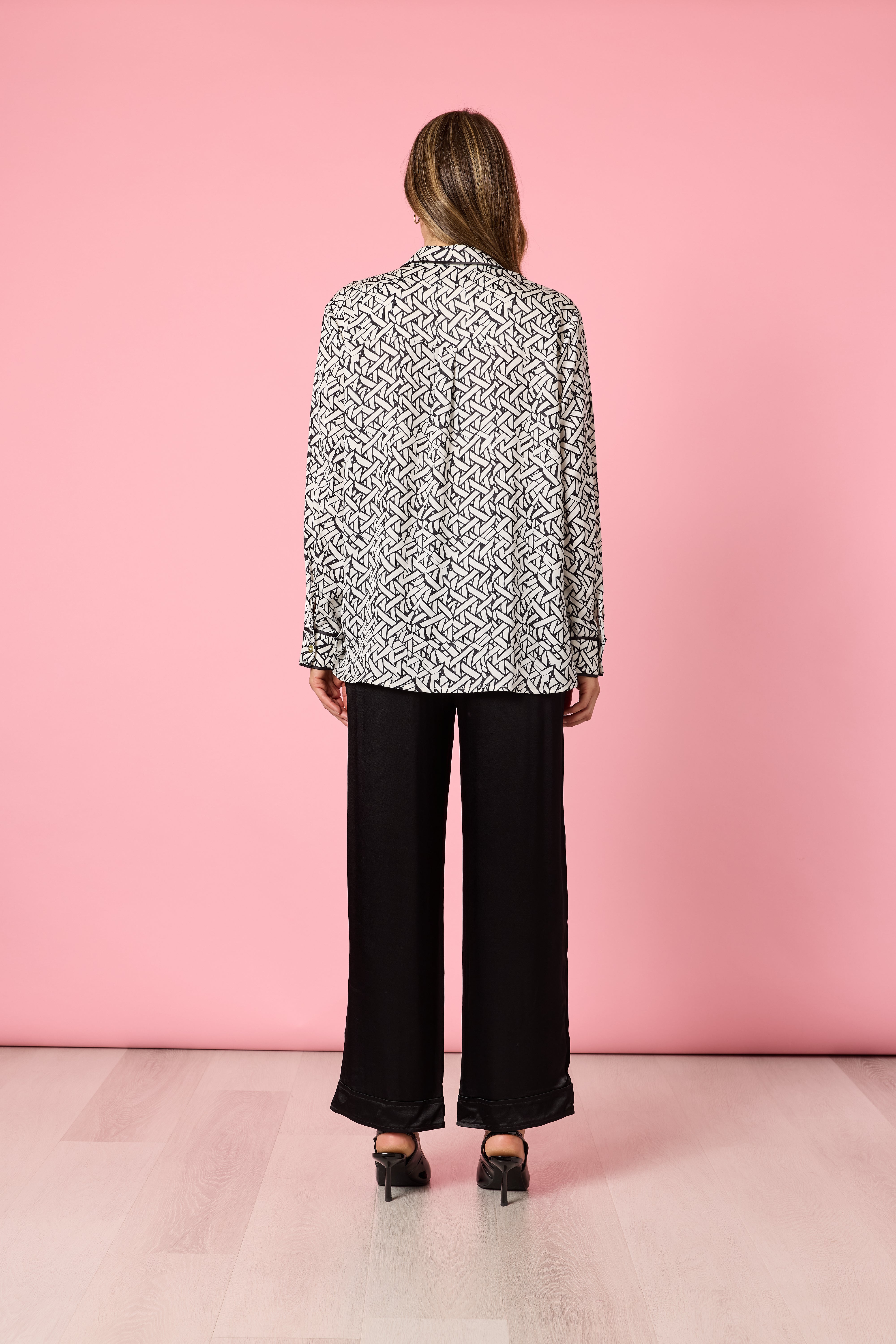 Back view of woman in black and ivory print blouse with loose black pants on pink background