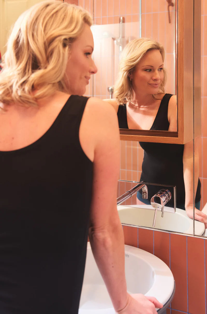 Woman wearing a black scoop neck bodysuit looking in bathroom mirror near sink