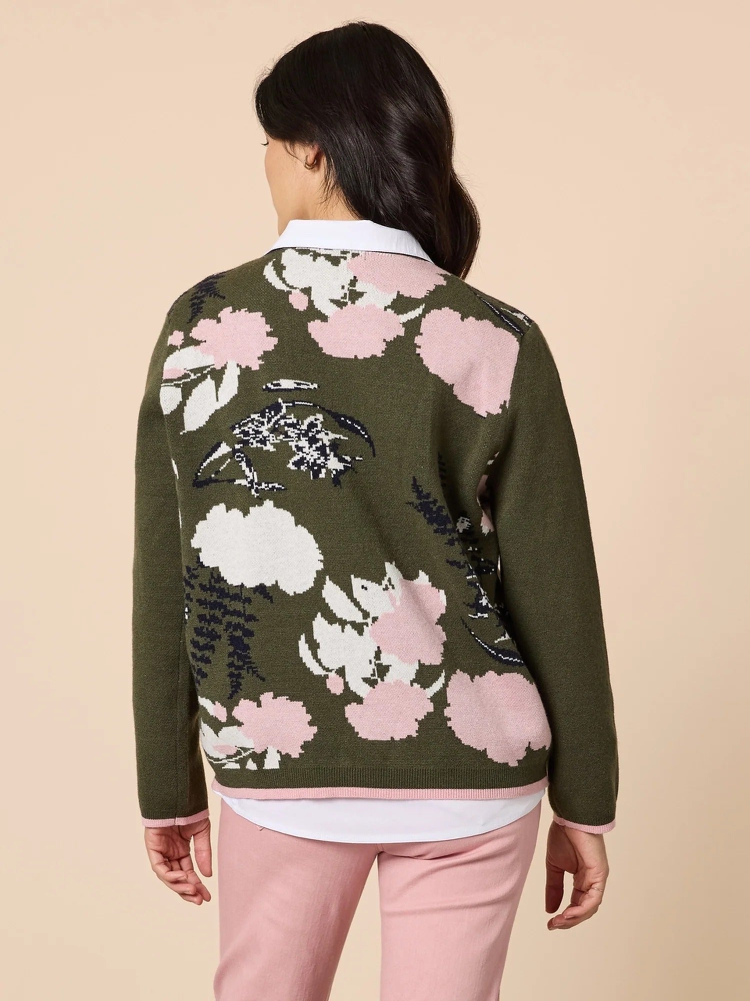 Back view of a woman wearing a khaki green floral knit sweater with pink and white patterns, paired with light pink pants against a beige background