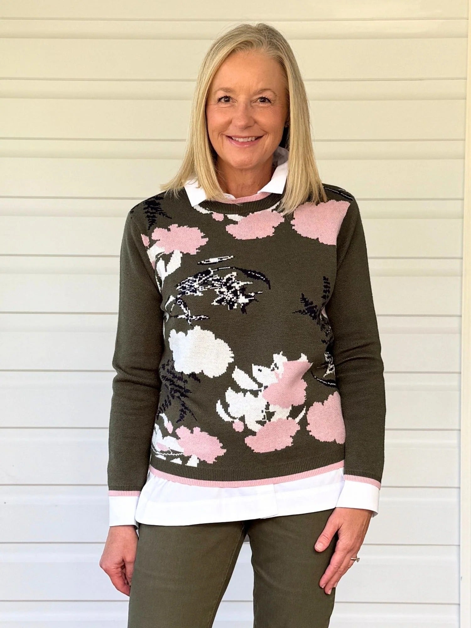Woman wearing a khaki green floral knit sweater with pink and white patterns layered over a white collared shirt, paired with matching khaki pants, standing against a light neutral background
