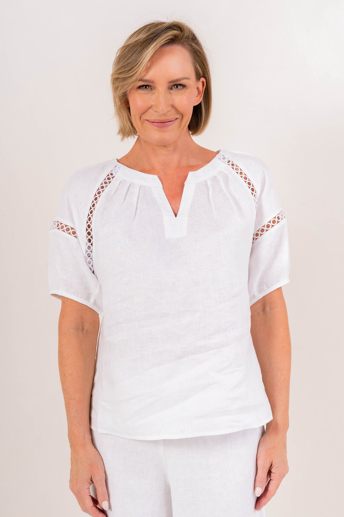 White linen embroidered top featuring a V-neckline, delicate cutwork detailing, short sleeves, and a relaxed silhouette for elegant everyday styling.
